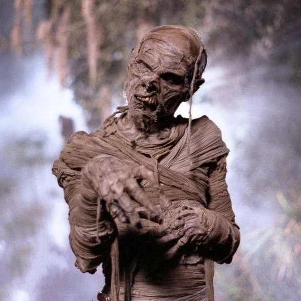 Create a dramatic image of a classic horror-style Mummy emerging from a shadowy, ancient Egyptian tomb, wrapped in aged linen bandages with glowing eyes and a haunting atmosphere.
